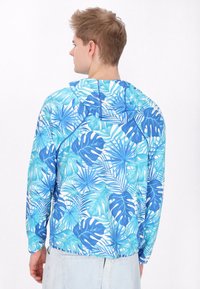 Pulover cu glugă, având un model de frunze tropicale în nuanțe de albastru și turcoaz, realizat din material ușor, cu mâneci lungi și o croială lejeră.