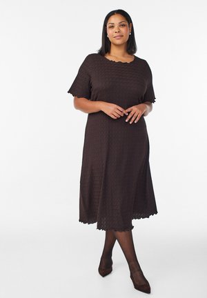 Kobieta stojąca w ciemnobrązowej, teksturowanej sukience midi z krótkimi rękawami i falistymi krawędziami, w przezroczystych rajstopach i ciemnych szpiczastych szpilkach na białym tle.