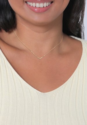 Goldene Halskette mit chevronförmigem Design, die am Halsansatz ruht, kombiniert mit einem leicht gerippten V-Ausschnitt-Oberteil. Glatte, metallische Textur.