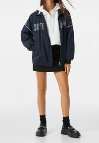 Marinblå bomberjacka med vit text, elastiska muddar och dragkedja fram. Buren över en vit hoodie, kombinerad med en svart minikjol och platåskor.