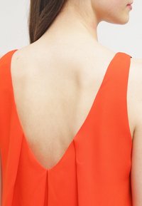 Červené šaty bez rukávů s hlubokým V-vyříznutím na zádech a měkkou, splývavou tkaninou. Materiál vypadá hladce s jemnými sklady.