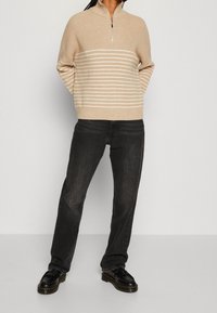 Béžový pletený svetr s límečkem na půl zip a vodorovnými bílými pruhy, v kombinaci s černými džínami a černými šněrovacími botami.
