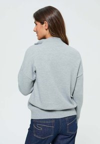 Pull gris avec un col haut, coupe ample et poignets côtelés, associé à un jean en denim sombre avec de subtils détails de couture.