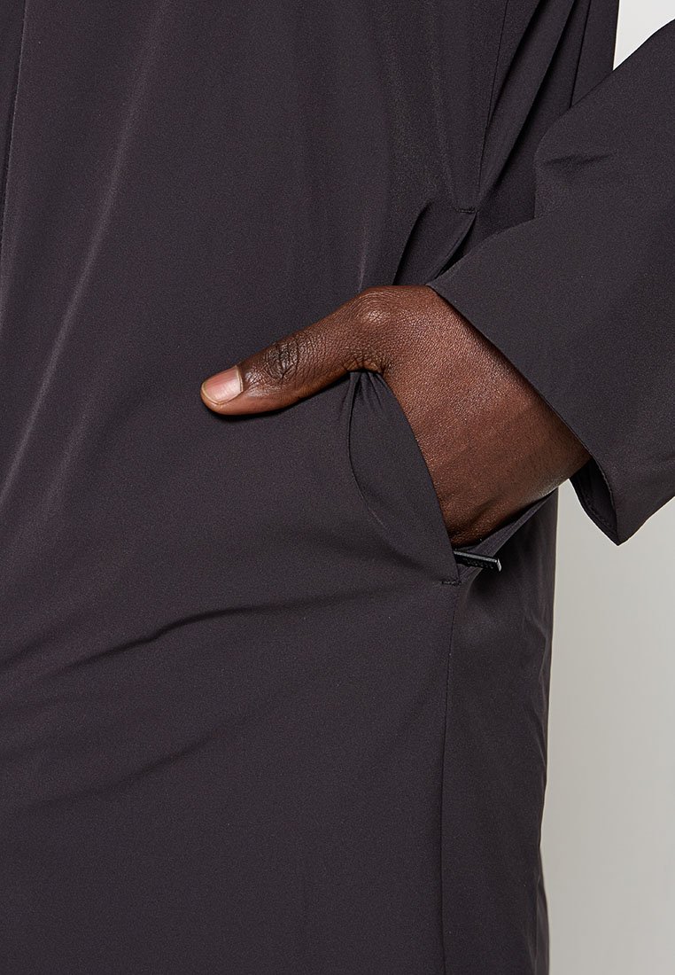 Czarna, gładka kurtka o gładkiej fakturze z boczną kieszenią. Materiał wydaje się lekkim i wodoodpornym. Ręka częściowo widoczna, bez wzorów ani elementów metalowych.