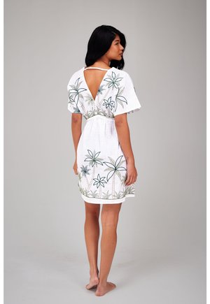 Femme aux longs cheveux foncés, debout pieds nus, portant une robe blanche avec une broderie de palmier et une découpe en V profonde dans le dos.