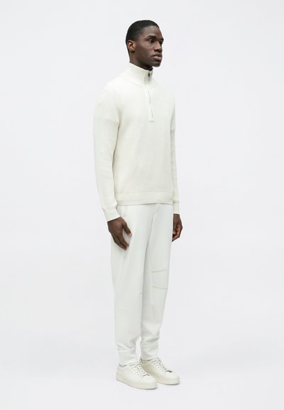 Pull-over en maille côtelée blanche avec col montant et fermeture éclair à moitié, associé à un pantalon blanc fuselé assorti et des baskets blanches.