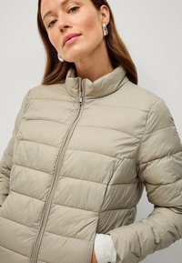 Abrigo puffer de color beige claro con patrón de acolchado horizontal, cuello alto y cremallera frontal. La tela parece suave y ligeramente brillante.