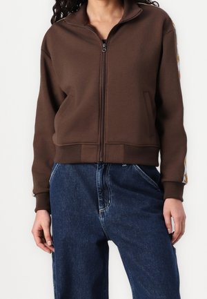 Femme portant une veste marron zippée avec des rayures à motifs sur les manches et un jean bleu ample taille haute.