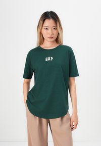 Kvinne med langt hår iført en mørkegrønn GAP t-skjorte og beige bukser, stående mot en enkel hvit bakgrunn.