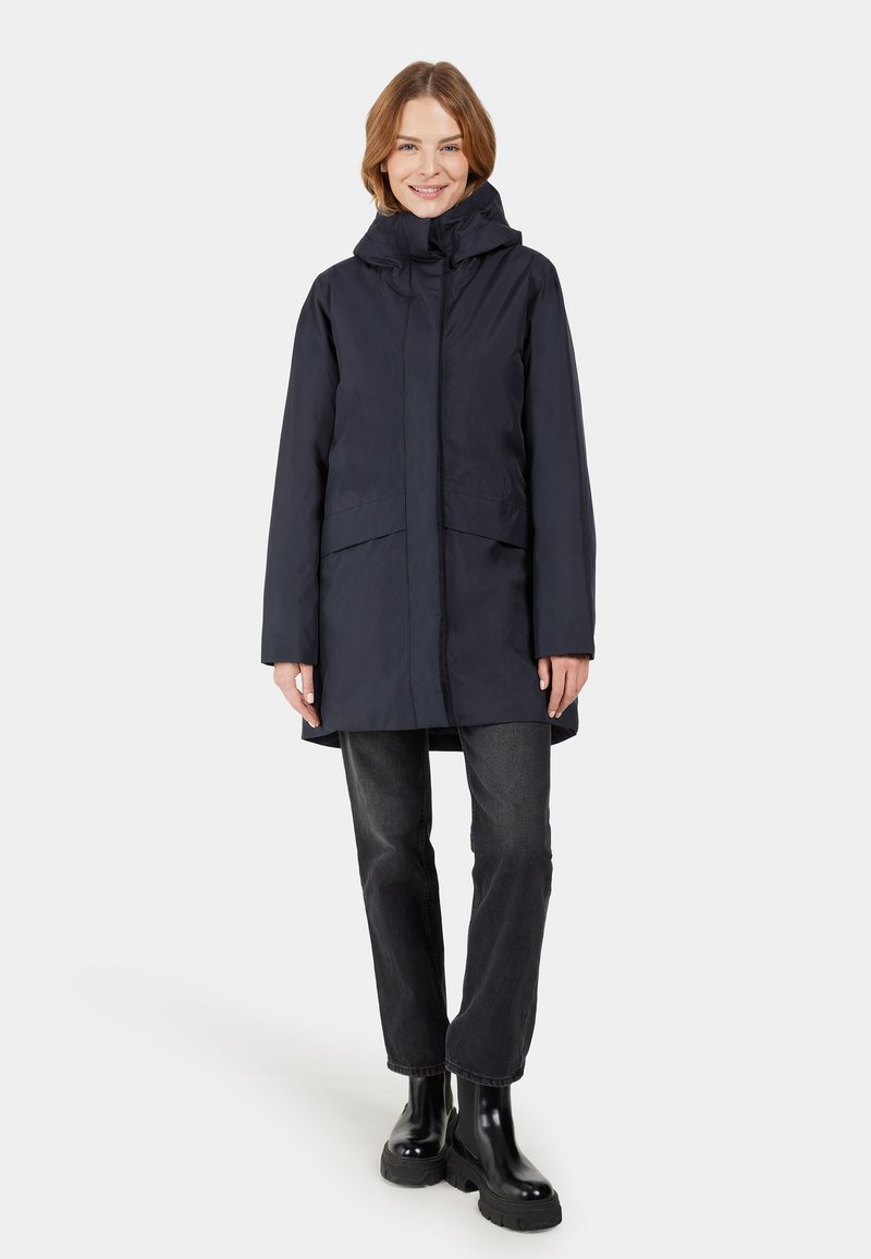 Marineblaue wasserdichte Parka mit Kapuze, zwei Fronttaschen und lockerer Passform, getragen über schwarzen Jeans und schwarzen Knöchelstiefeln.