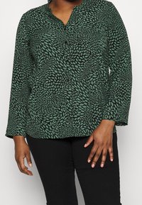 Blusa verde con motivo a foglia, maniche lunghe, chiusura con bottoni e scollatura a V. Il tessuto ha una consistenza morbida con una leggera lucentezza.