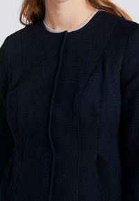 Granatowy płaszcz wełniany z domieszką, z okrągłym dekoltem, wyraźnymi szwami i dwoma bocznymi kieszeniami, z kontrastującym jasnoszarym wewnętrznym kołnierzem.