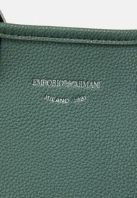 Zelená hnětá kůže se stříbrným logem "Emporio Armani" a detailní embosovanou značkou "Milano 1981" pod ním. Hladký povrch s jemným leskem.