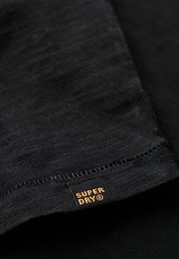 Zwarte stoffen t-shirt met een soepele textuur. Bevat een klein, gouden 'SUPERDRY' label nabij de zoom, met een eenvoudige gestikte rand.