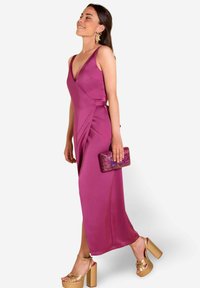 Vestido de satén magenta con escote pronunciado y diseño envolvente, combinado con un bolso de mano púrpura brillante y sandalias de tacón bloque doradas.