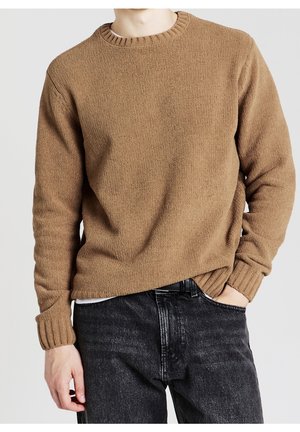 Homme portant un pull en maille marron avec poignets et col côtelés, associé à un jean noir délavé, main dans la poche, sur un fond uni.