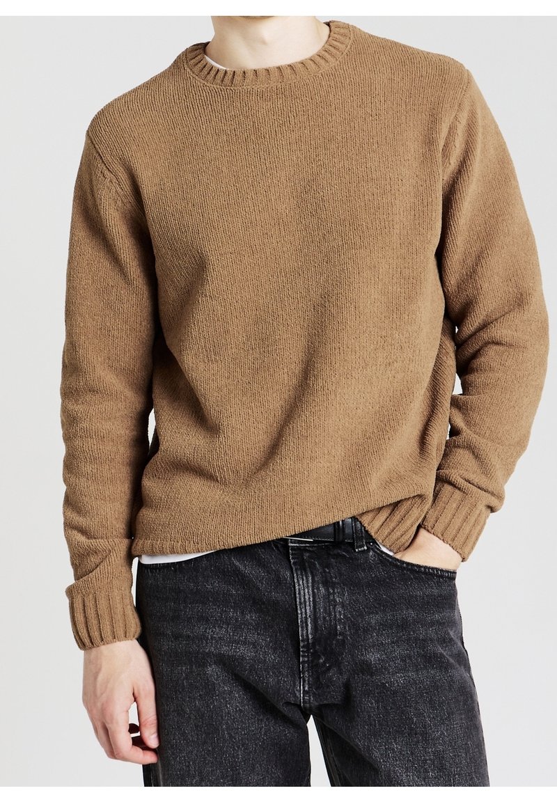 Homme portant un pull en maille marron avec poignets et col côtelés, associé à un jean noir délavé, main dans la poche, sur un fond uni.