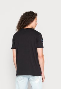 Čierne tričko s krátkym rukávom s okrúhlym výstrihom a farebnou nášivkou na ľavom pleci. Vyrobené z mäkkej, štruktúrovanej bavlny.