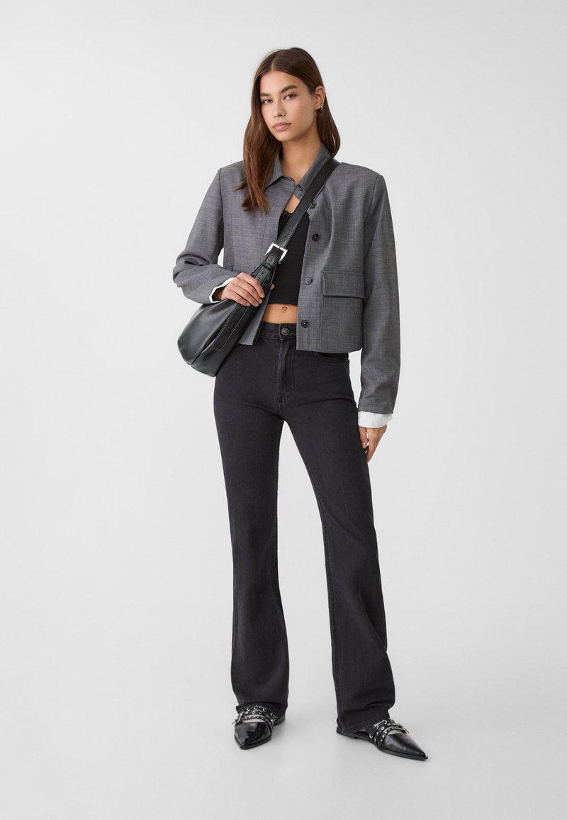 Femme jeune portant un blazer gris court sur un haut noir court, un jean noir évasé, des chaussures noires pointues et portant un sac à bandoulière noir.
