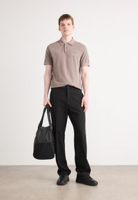Brun polo skjorte med korte ermer, svarte bukser og svarte sneakers. Holder en svart bag med teksturert overflate. Minimalistisk design.
