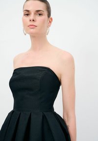 Utan axelband svart klänning med texturerad tyg, figursydd överdel och utsvängd kjol. Modellen har stora metallic-hoopörhängen. Bakgrunden är helt vit.