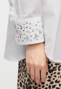 Μανσέτα λευκού πουκαμίσου διακοσμημένη με ροζ και διαυγή πολύτιμους λίθους, με λεία υφή και λεπτομέρεια με κουμπί. Φόντο με μοτίβο λεοπάρδαλης.