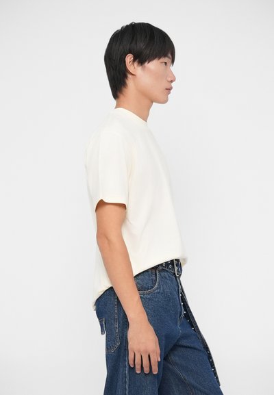 Camiseta de manga corta crema con cuello redondo, combinada con unos vaqueros de denim azul de cintura alta que cuentan con un bolsillo lateral y detalles de botones.