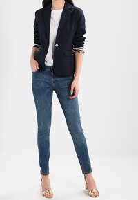 Granatowa marynarka z pojedynczym guzikiem i klapami, sparowana z białą koszulką i niebieskimi obcisłymi dżinsami. Złote sandały na obcasie dopełniają stylizację.