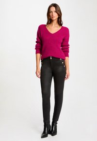 Pull en tricot fuchsia avec un col en V et des manches larges, associé à un jean skinny noir avec des accents de boutons sur le côté et des bottes à talons.