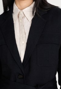 Žena má na sobě béžovou košili na knoflíky pod černým sakem s prsu kapes a jedním viditelným knoflíkem zapnutým.