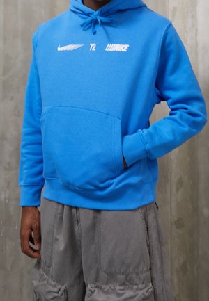 Personne portant un sweat à capuche Nike bleu vif avec un logo blanc et le numéro 72, assorti à un pantalon cargo gris ample, debout contre un mur uni.