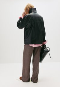 Schwarze Kunstlederjacke mit hohem Kragen, über einem rosa-weiß gestreiften Hemd getragen, kombiniert mit hellbraunen, weiten Hosen.