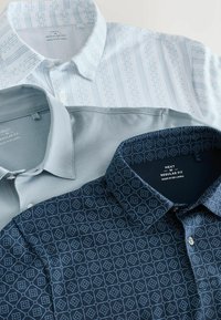 Tres camisas de cuello: una blanca con rayas verticales azules, una lisa azul claro y una azul marino con patrón de rombos. Todas tienen una textura suave.