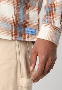 Brown and blue plaid flannel shirt with cream background, featuring a blue label. Cream-colored fabric pants with a side pocket.