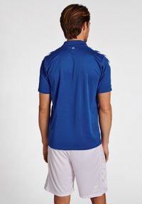 Blå polo shirt med en glat tekstur, prydet med hvide detaljer på skuldrene, sammen med hvide shorts. Set bagfra.