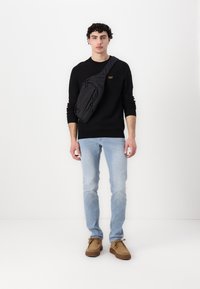 Zwarte lange mouw shirt, lichtblauwe jeans, bruine schoenen. Zwart schoudertasje met ritsen. Effen achtergrond benadrukt de outfit.