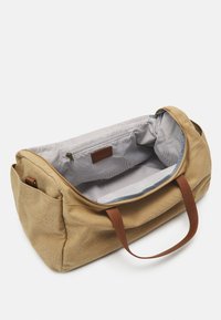 Beige kankaan duffel-laukku ruskeilla nahkakahvoilla. Sisäpuolella on raidallinen kuvio ja vetoketjullinen tasku. Avoin muotoilu helpottaa pääsyä.