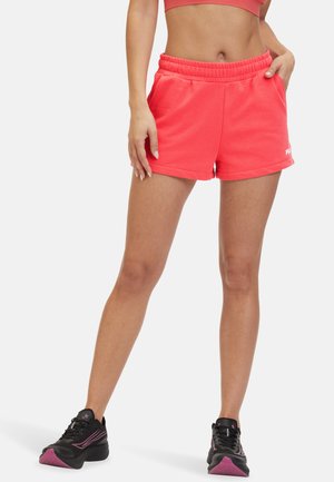 Pantaloni sportivi rosa in cotone con vita elasticizzata, tasche laterali e un dettaglio logo bianco. Indossati con scarpe da ginnastica nere.