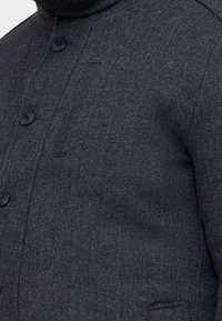 Dunkelgrauer Fischgrätenjacke mit hohem Kragen, Knopfverschluss und strukturiertem Stoff. Enthält eine Brusttasche und eine glatte Oberfläche.