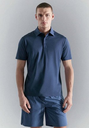 Giovane uomo in piedi davanti a uno sfondo semplice, indossa una polo blu navy a maniche corte e pantaloncini abbinati.