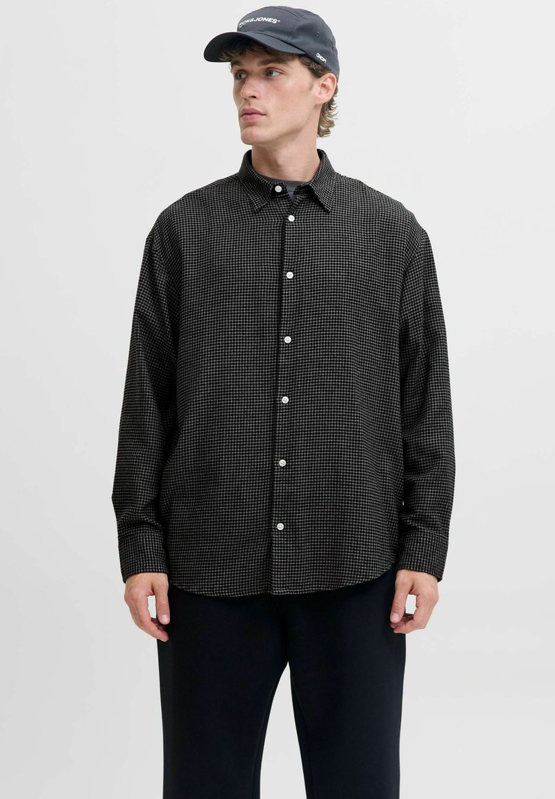 Camisa de botones a cuadros en blanco y negro con mangas largas, cuello y botones blancos. Llevada con pantalones oscuros y una gorra oscura.