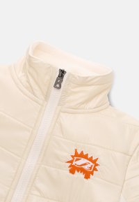 Pudret, cremefarvet jakke med høj krave, lynlåslukning og orange broderet logo. Har vandret quiltning og glat tekstur.