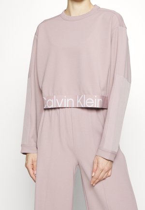 Frau trägt ein hellmaulbeerfarbiges, langärmeliges, kurzes Sweatshirt und passende, weit geschnittene Hosen mit einem sichtbaren Calvin Klein Logo-Band.