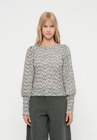 Γυναίκα με ίσια ξανθά μαλλιά, φορώντας μια γκρι υφασμάτινη μπλούζα με μακριά μανίκια και σκούρους γκρι παντελόνι σε φαρδιά γραμμή, που στέκεται μπροστά σε λευκό φόντο.