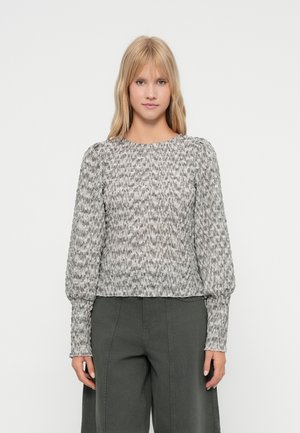 Γυναίκα με ίσια ξανθά μαλλιά, φορώντας μια γκρι υφασμάτινη μπλούζα με μακριά μανίκια και σκούρους γκρι παντελόνι σε φαρδιά γραμμή, που στέκεται μπροστά σε λευκό φόντο.