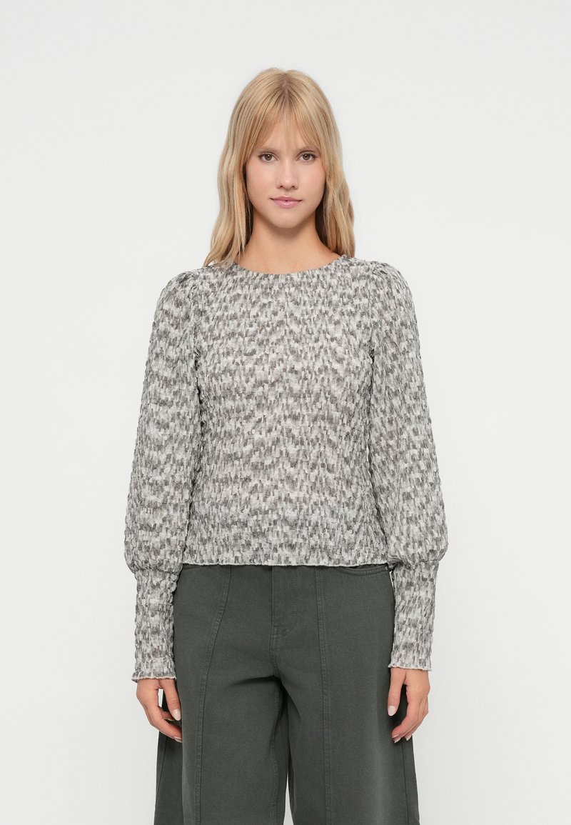 Γυναίκα με ίσια ξανθά μαλλιά, φορώντας μια γκρι υφασμάτινη μπλούζα με μακριά μανίκια και σκούρους γκρι παντελόνι σε φαρδιά γραμμή, που στέκεται μπροστά σε λευκό φόντο.