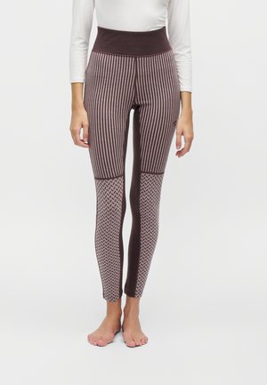 Femme portant des leggings taille haute avec des rayures verticales et des motifs géométriques en marron et beige, debout pieds nus devant un fond uni.
