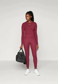 Maroon urheiluvaatesetti, joka on valmistettu joustavasta kankaasta. Pitkähi mittainen paita on yhdistetty korkeavyötäröisiin legginsseihin. Musta jumppakassi pidetään vasemmassa kädessä. Valkoiset lenkkarit.