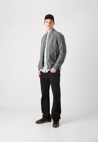 Pilkas bomber tipo švarkas, šviesiai mėlynas dryžuotas marškinėliai, juodos džinsai, juodi sportbačiai. Modelis stovi prieš neutralų foną.