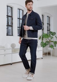 Donkerdenimjack over een lichtgrijze trui, gecombineerd met een donkere spijkerbroek en meerkleurige sneakers. Felle binnenomgeving met planten en meubels.